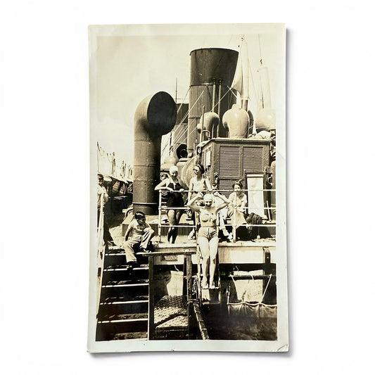 Vintage black and white photograph of people on a ship's deck with visible ship equipment. A woman getting ready to jump into a pool.