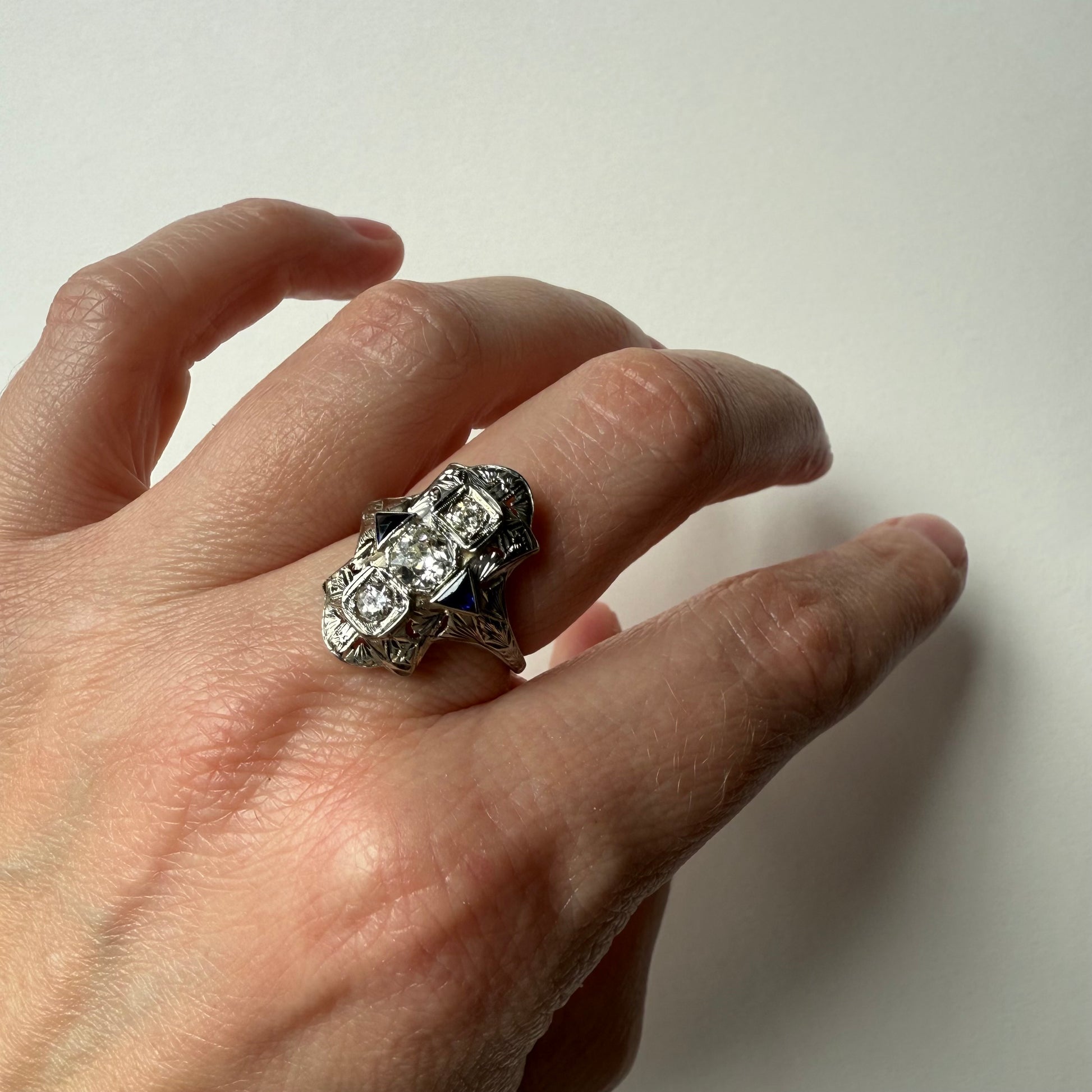 Hand wearing an art deco 18kt white gold cocktail engagement ring with diamonds and sapphires on a plain background