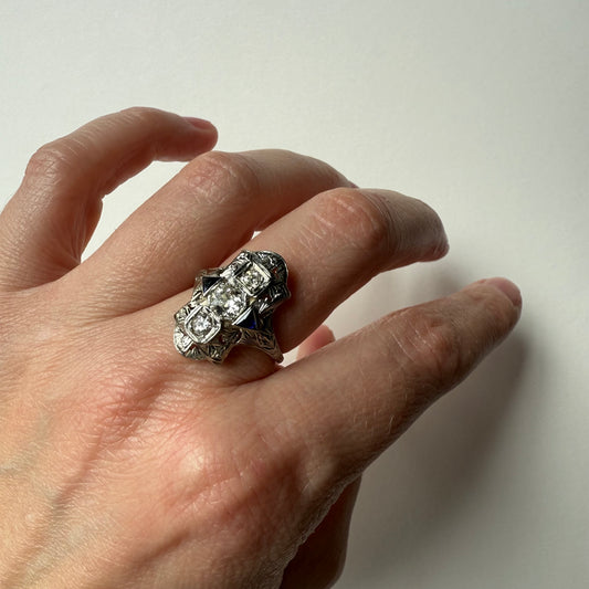 Hand wearing an art deco 18kt white gold cocktail engagement ring with diamonds and sapphires on a plain background