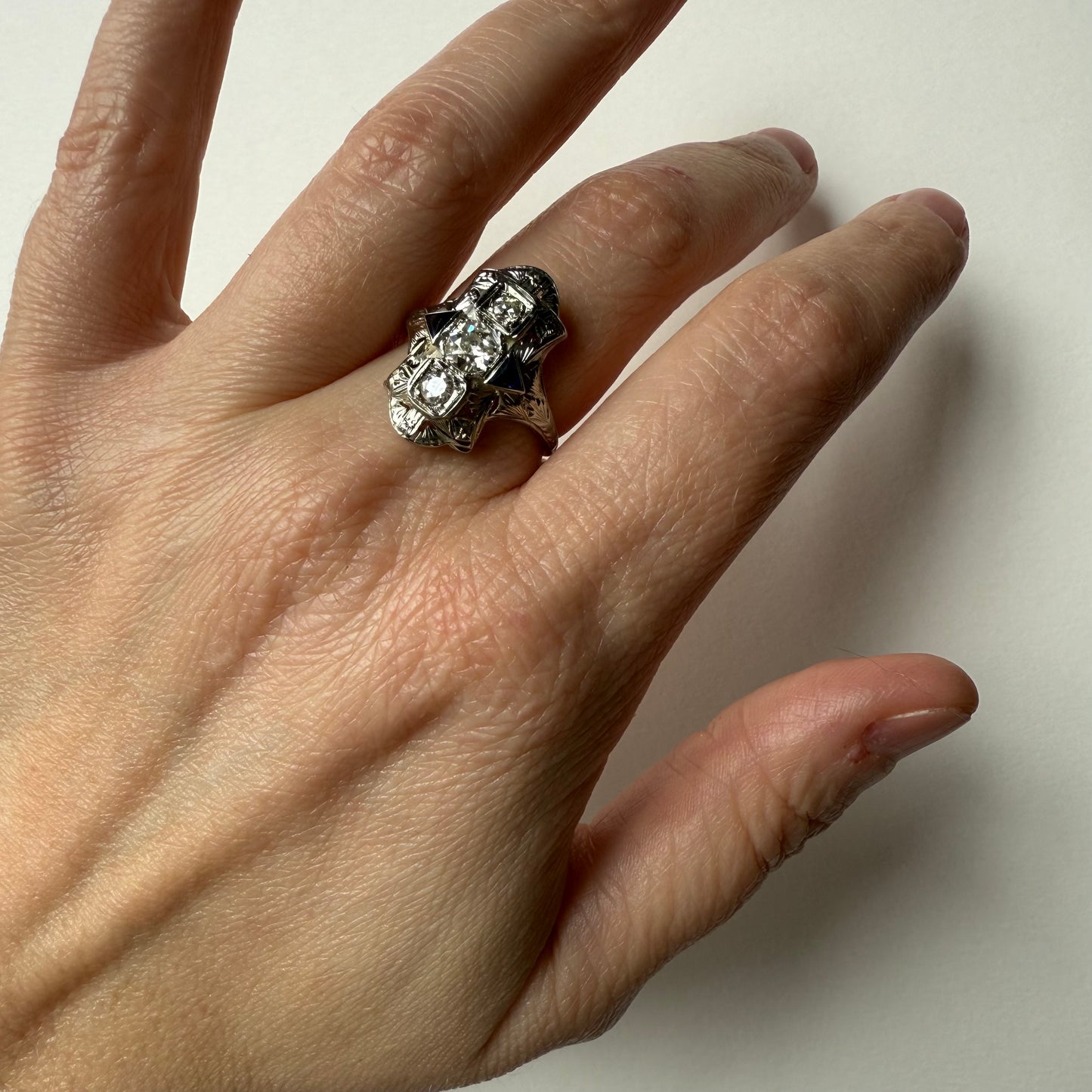 Hand wearing an art deco 18kt white gold cocktail engagement ring with diamonds and sapphires on a plain background