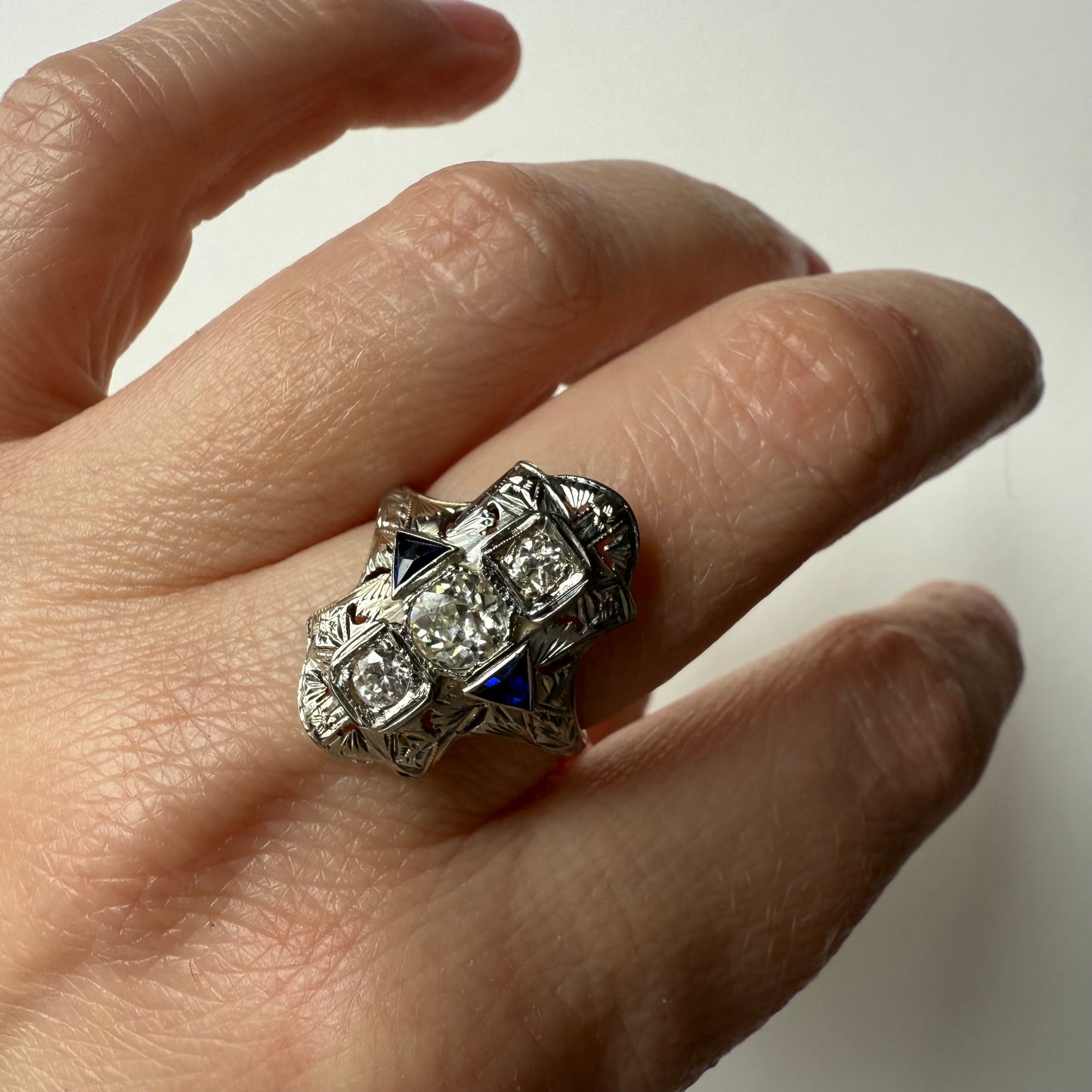 close- up of Hand wearing an art deco 18kt white gold cocktail engagement ring with diamonds and sapphires on a plain background
