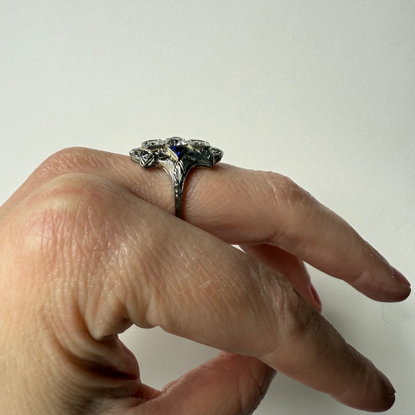 Hand wearing an art deco 18kt white gold cocktail engagement ring with diamonds and sapphires on a plain background