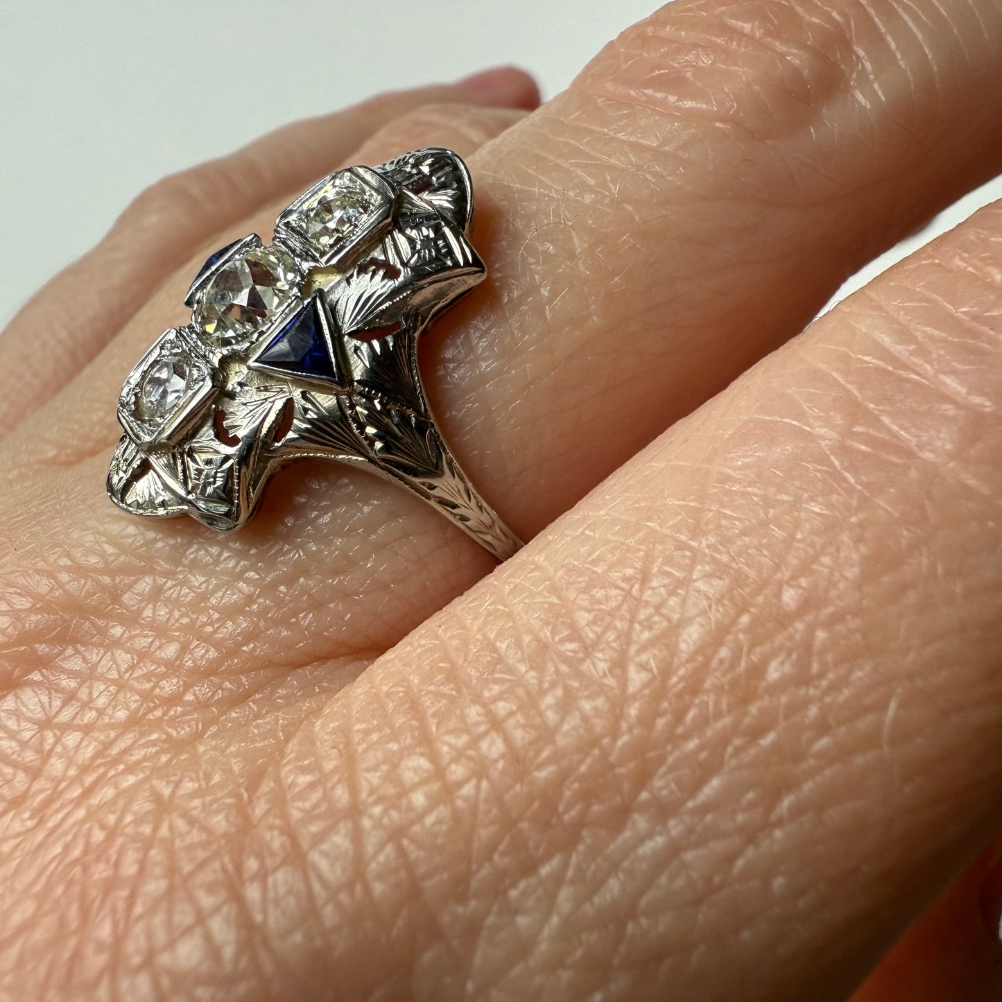 Hand wearing an art deco 18kt white gold cocktail engagement ring with diamonds and sapphires on a plain background
