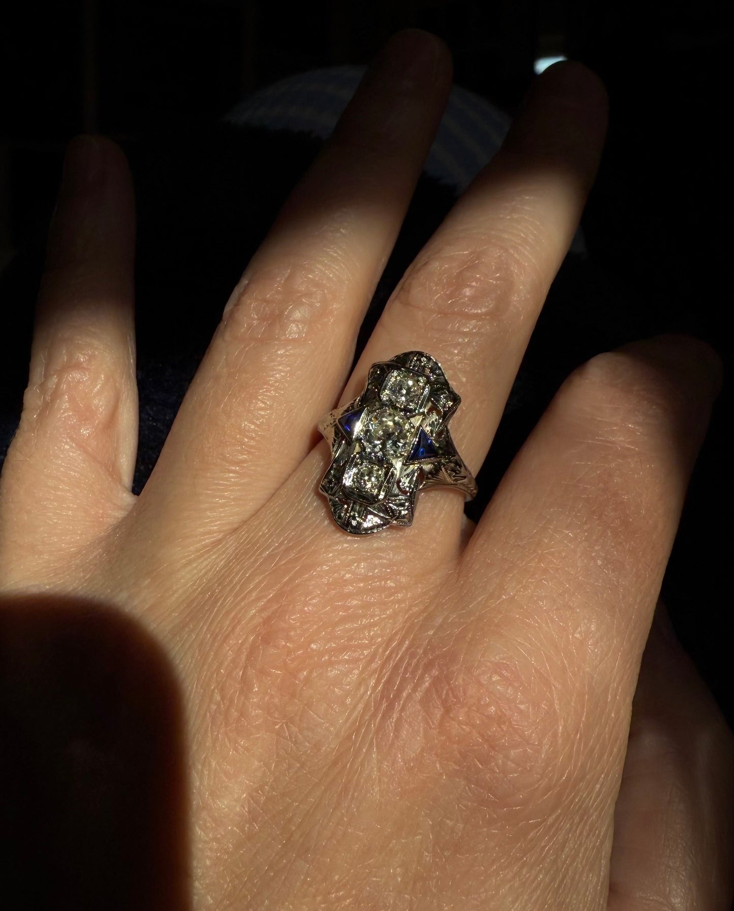 Hand wearing an art deco cocktail engagement ring with diamonds and sapphires on a plain background