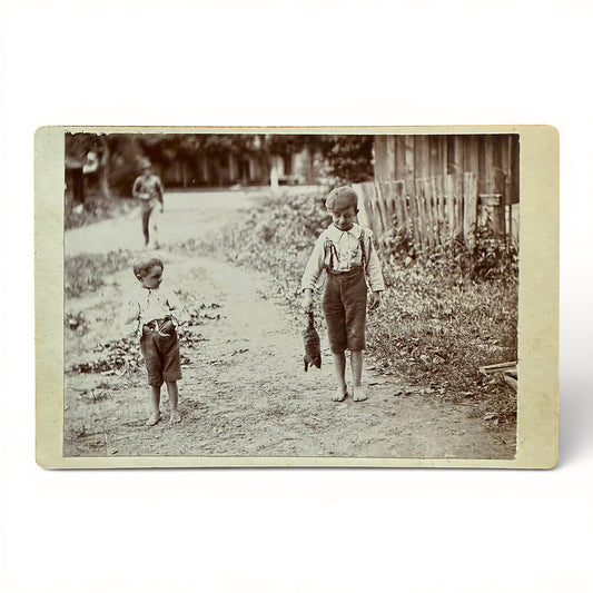 Caught A New Pet - Cabinet Card Vintage black and white photo of two children walking on a dirt path with a fence and trees in the background. one kid has caught a squirrel.