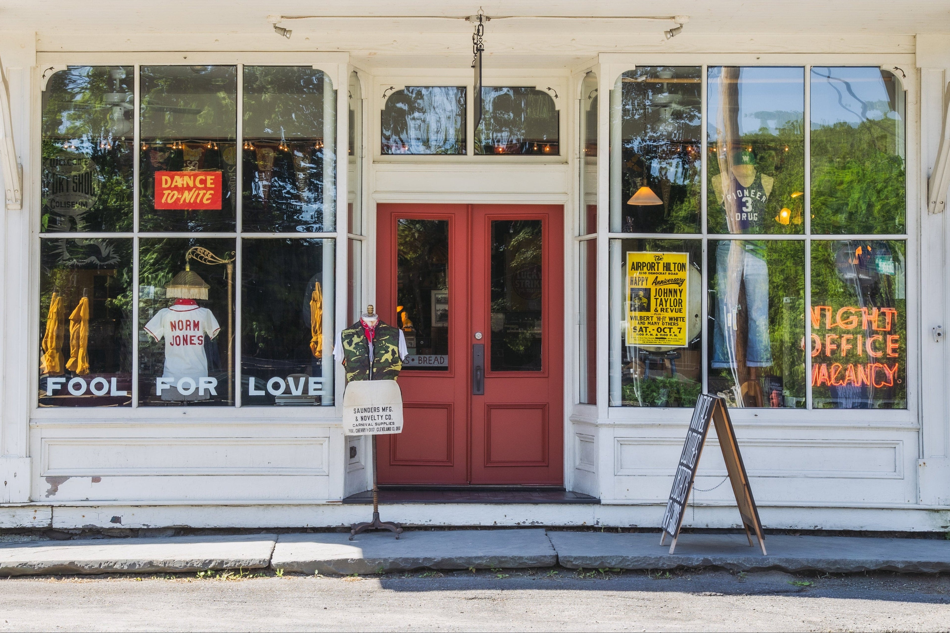 Two-story building with a red door and glass windows with an antique neon sign and window display on a street corner. A vintage store retail location.