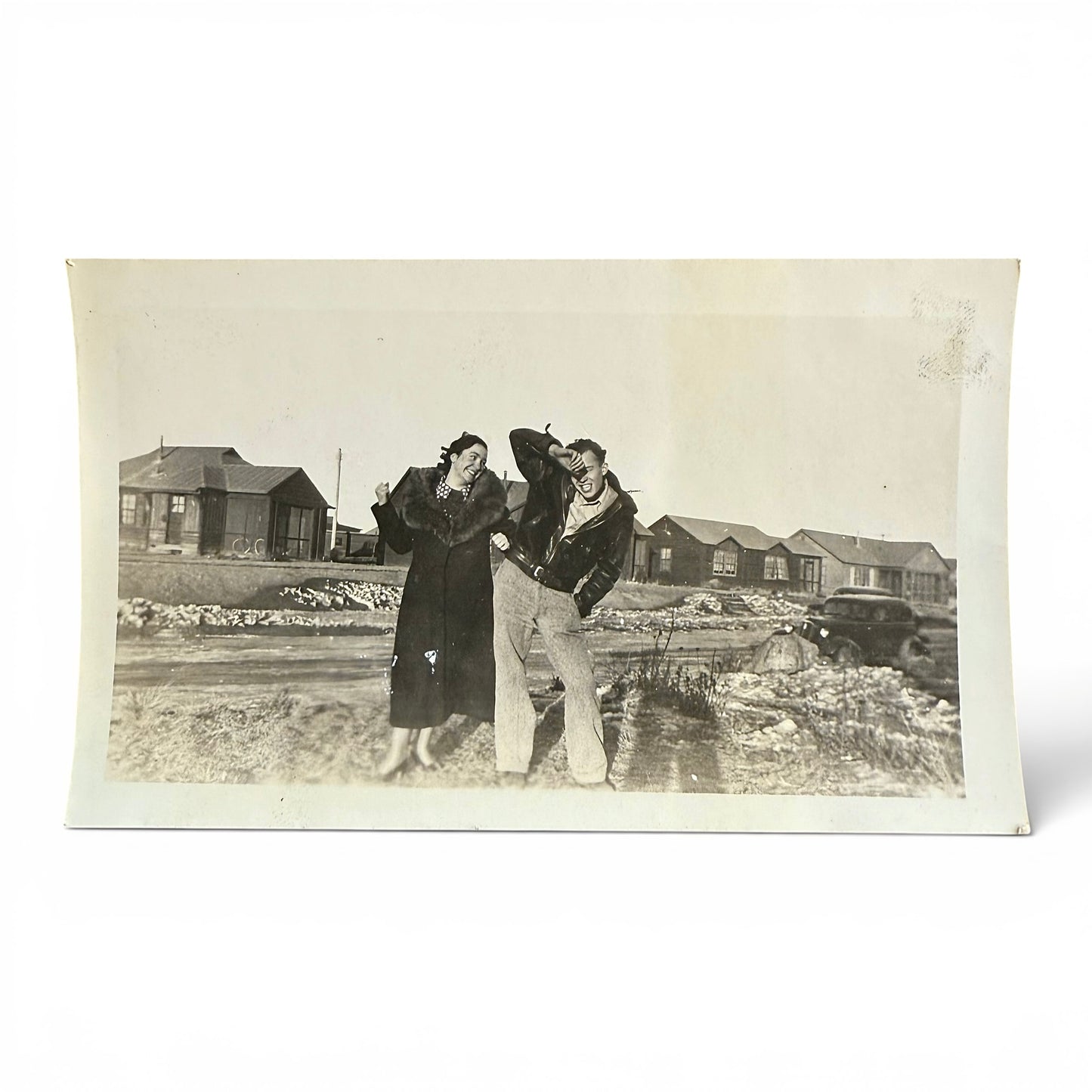 Vintage black and white photo of two people being playful and standing outdoors with small buildings in the background.