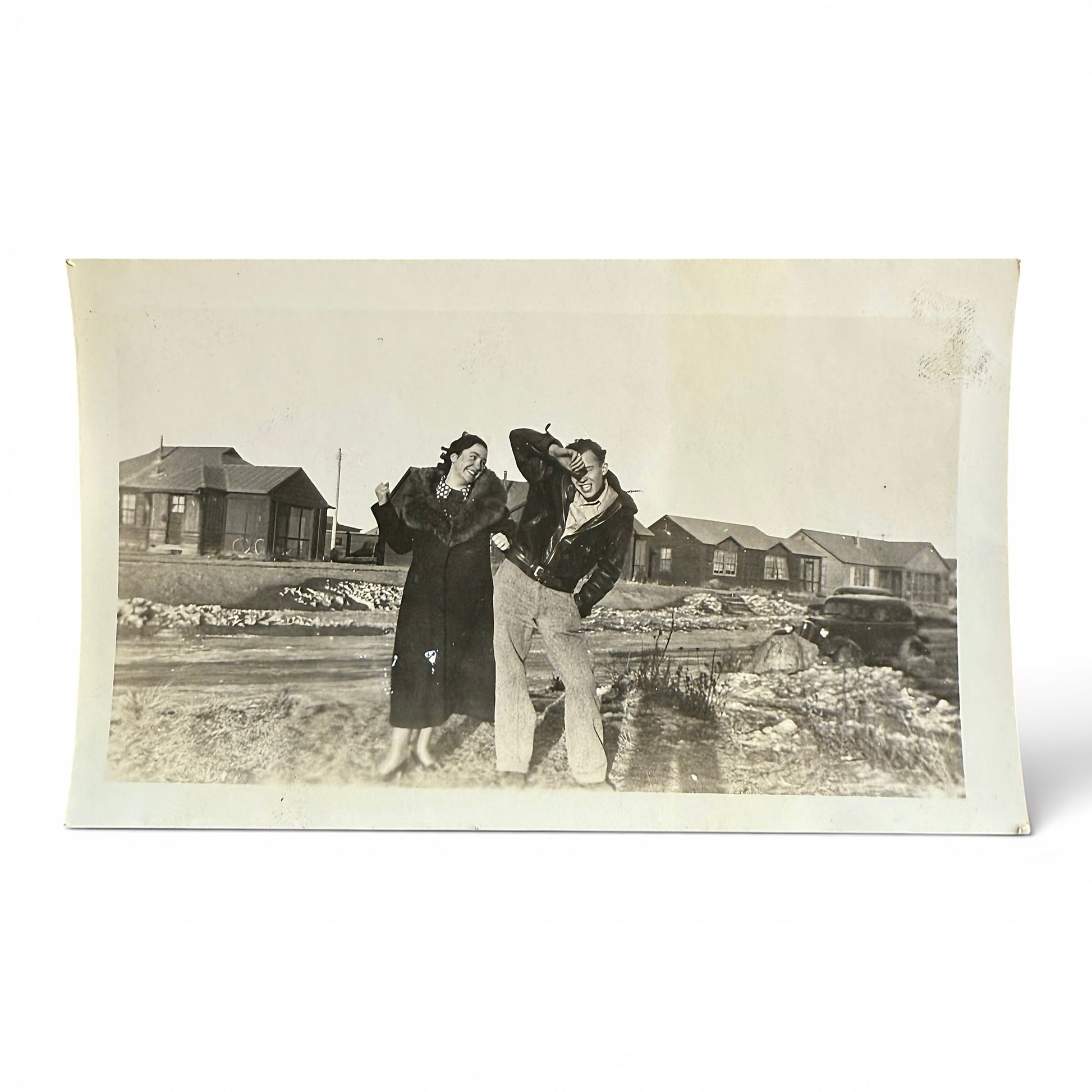 Vintage black and white photo of two people being playful and standing outdoors with small buildings in the background.